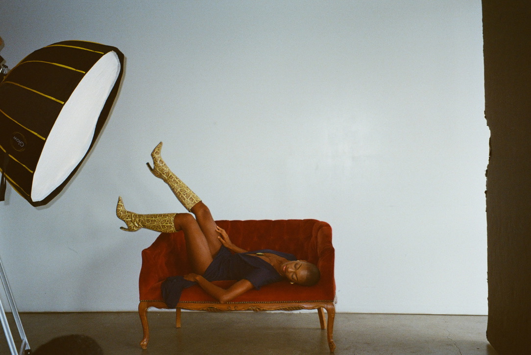 Model in a navy dress and gold boots reclining on a red velvet settee